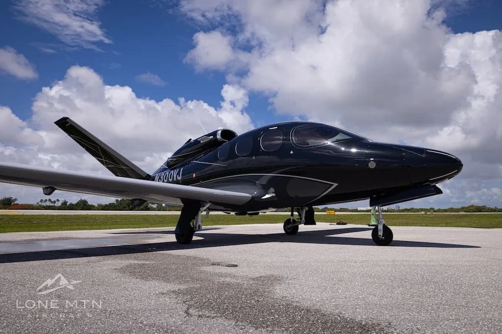 A black Cirrus Vision Jet (N300VJ) with silver accents parked on an airport tarmac under a blue sky with clouds, viewed from the front-right.
