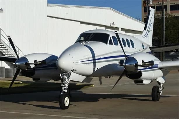 A white twin-engine turboprop aircraft with blue and grey stripes is parked on an apron in front of a white hangar.