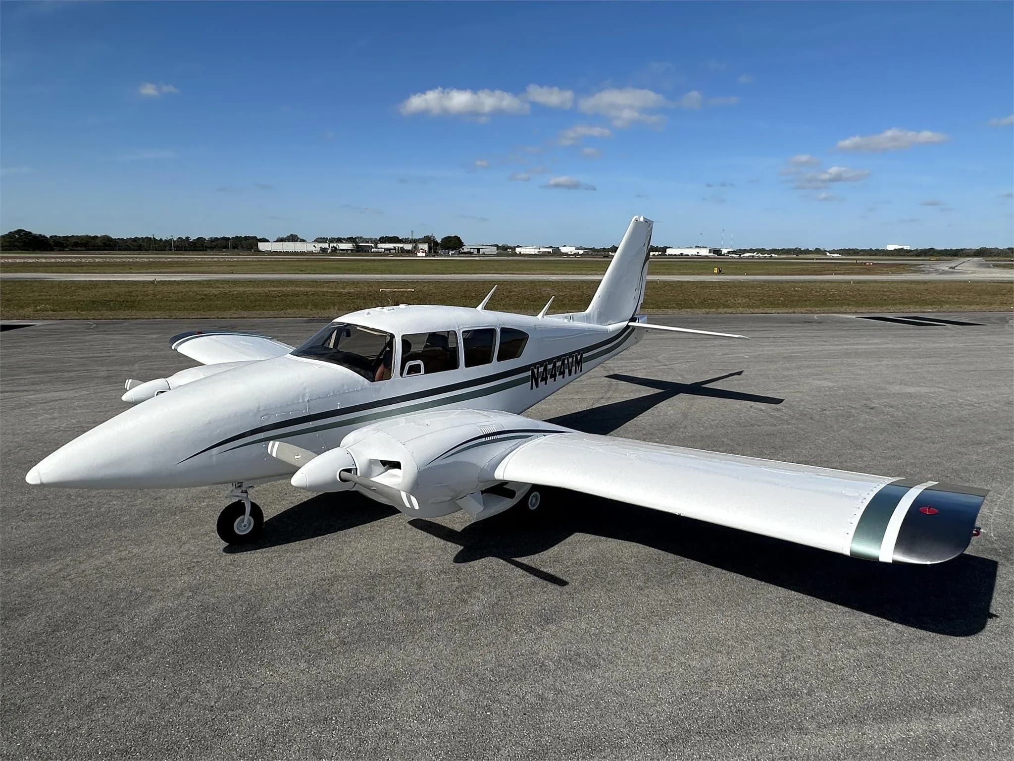 A white twin-engine propeller aircraft with green stripes on the fuselage and wingtips, parked on an asphalt runway under a blue sky.