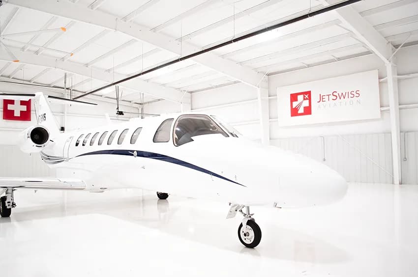 A white business jet with blue stripes (N716NS) parked in a brightly lit hangar, viewed from the front-right.