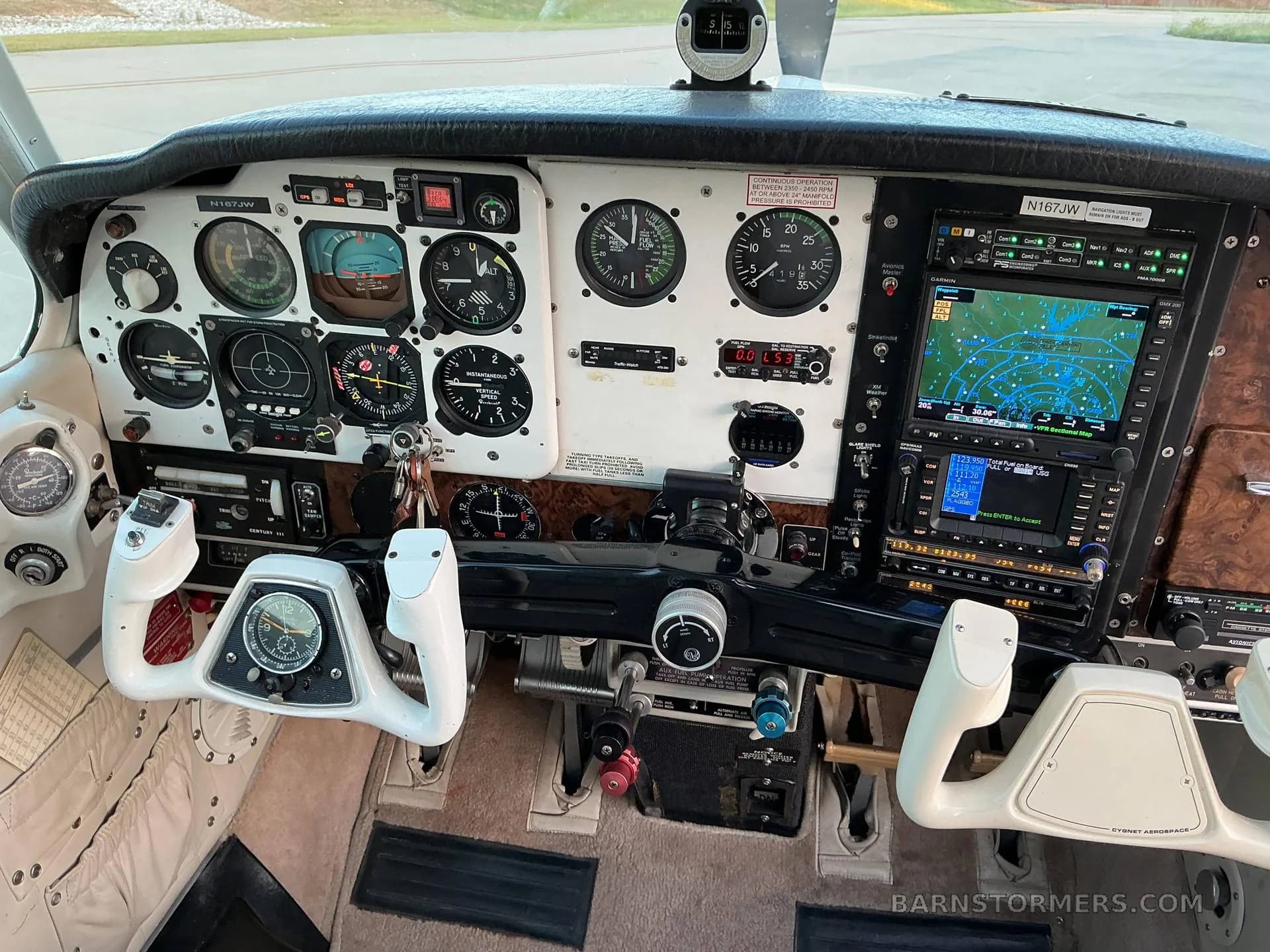The cockpit of a Beechcraft Bonanza (N167JW) showing the instrument panel with analog gauges, a modern GPS display, and white control yokes.