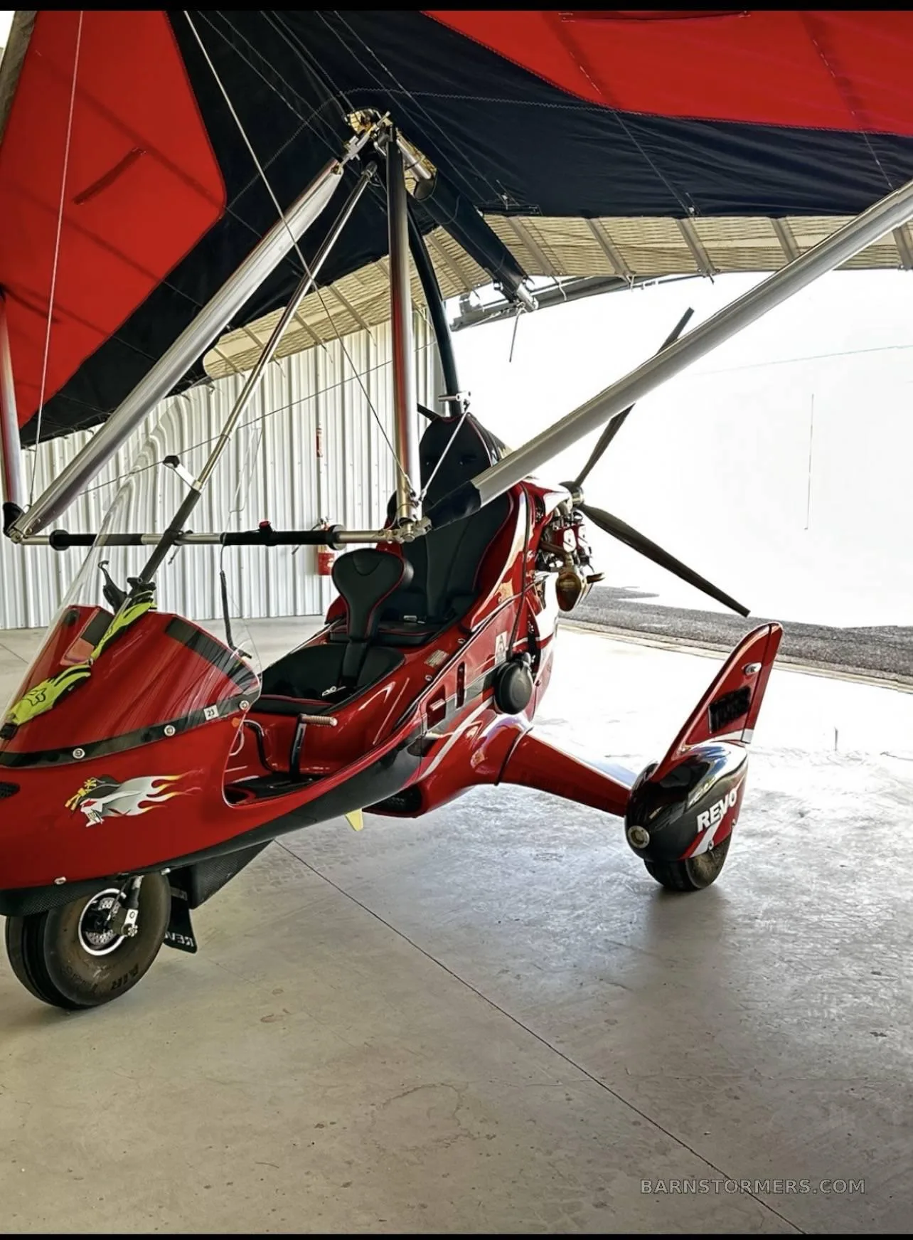 A red weight-shift control aircraft with a black and red wing is parked inside a hangar.