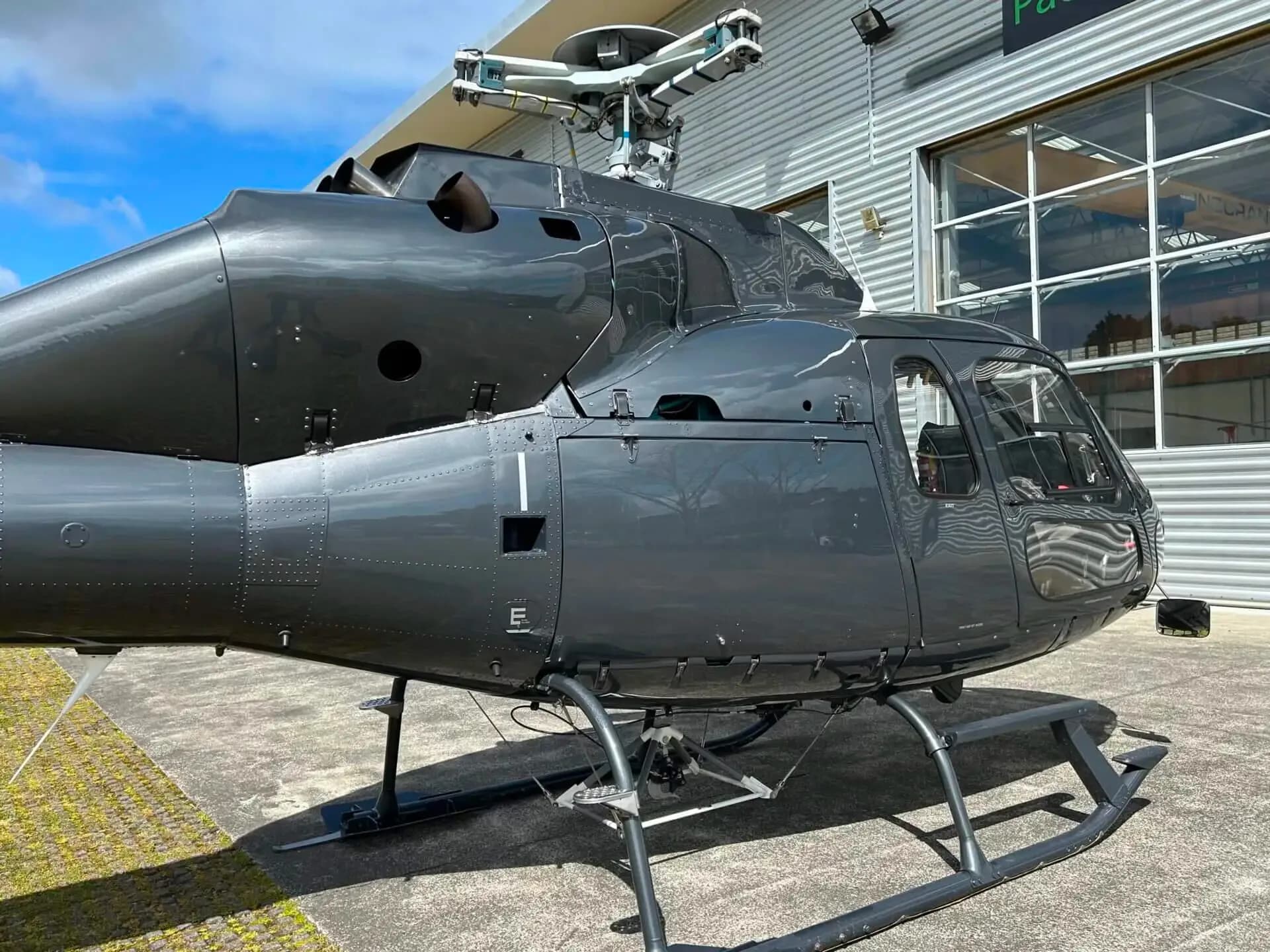 A side view of a dark grey Eurocopter AS355 F1 helicopter, showing its fuselage and landing skids, parked outside a hangar.