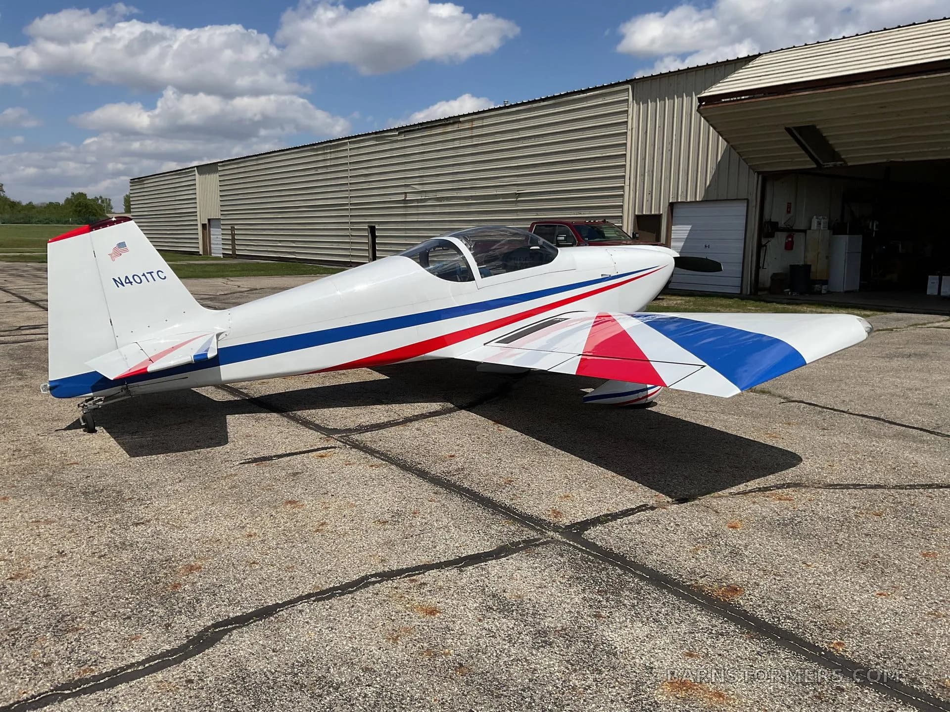 A white single-engine aircraft with red and blue stripes on its wings and tail (N401TC), parked on an asphalt tarmac with a hangar in the background.