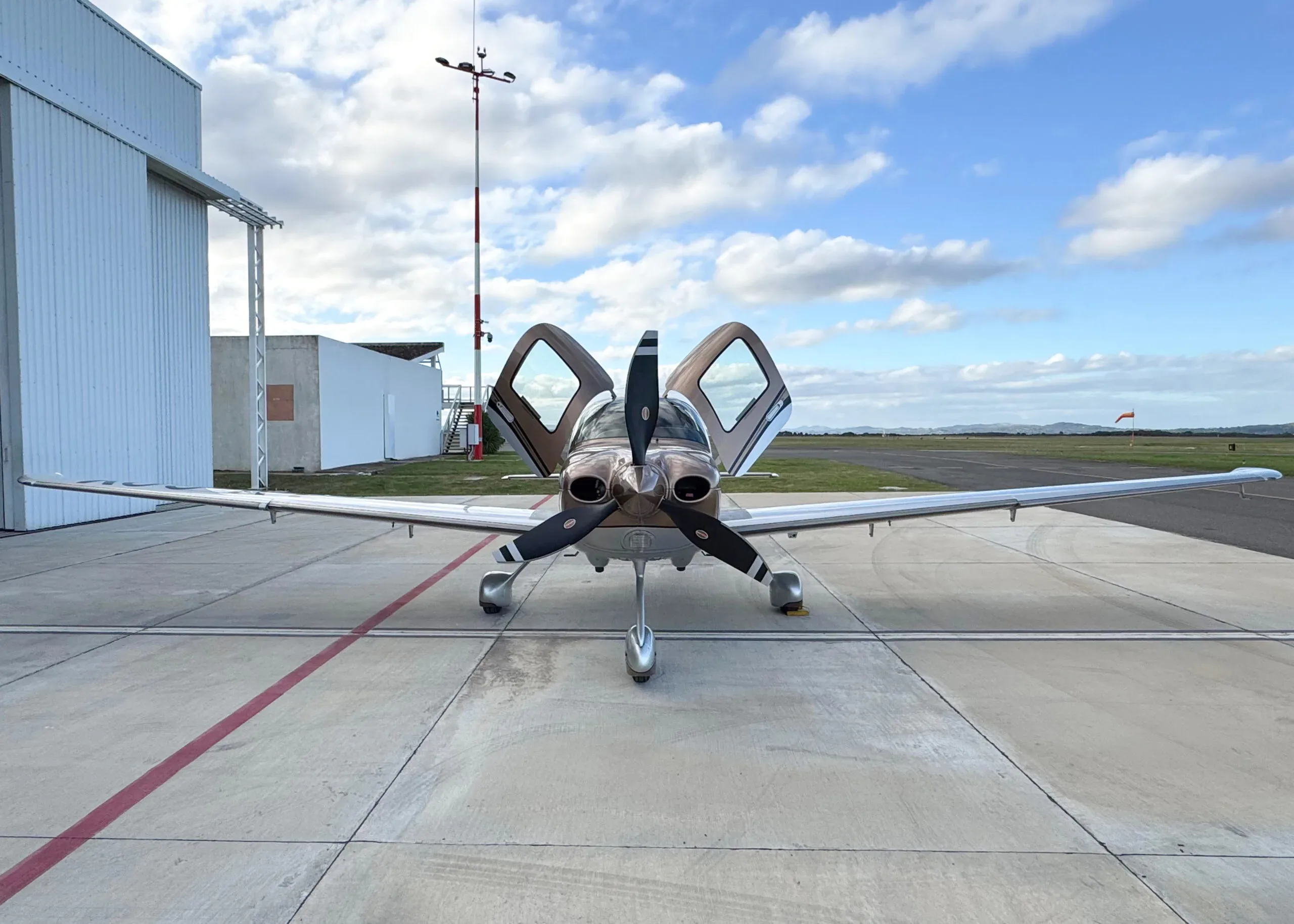 A front view of a brown and silver Cirrus SR22T aircraft with its gull-wing doors open, parked on the tarmac.
