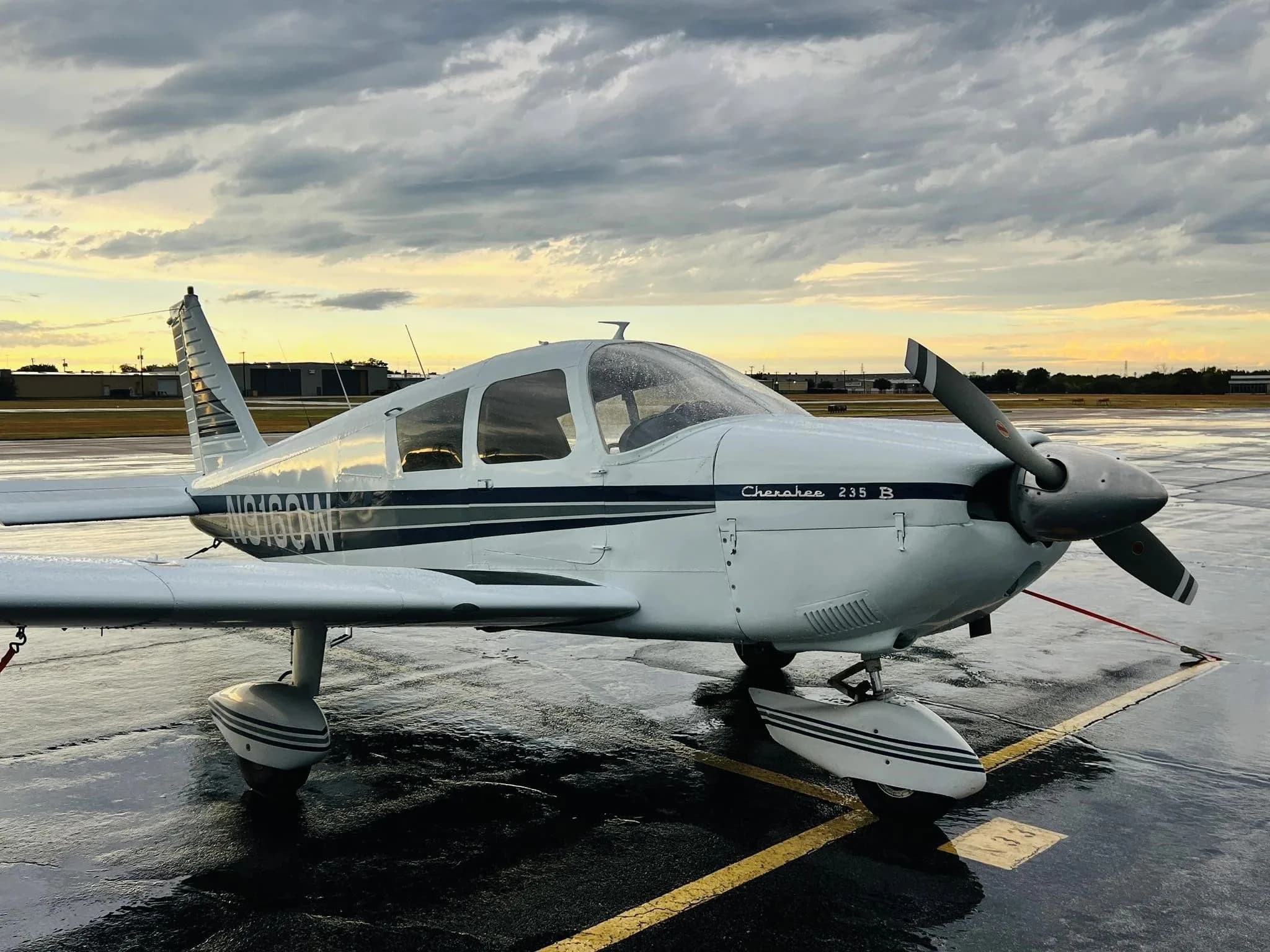 Front-side view of a white Piper Cherokee 235 B (N9160W) on a wet tarmac at sunset.