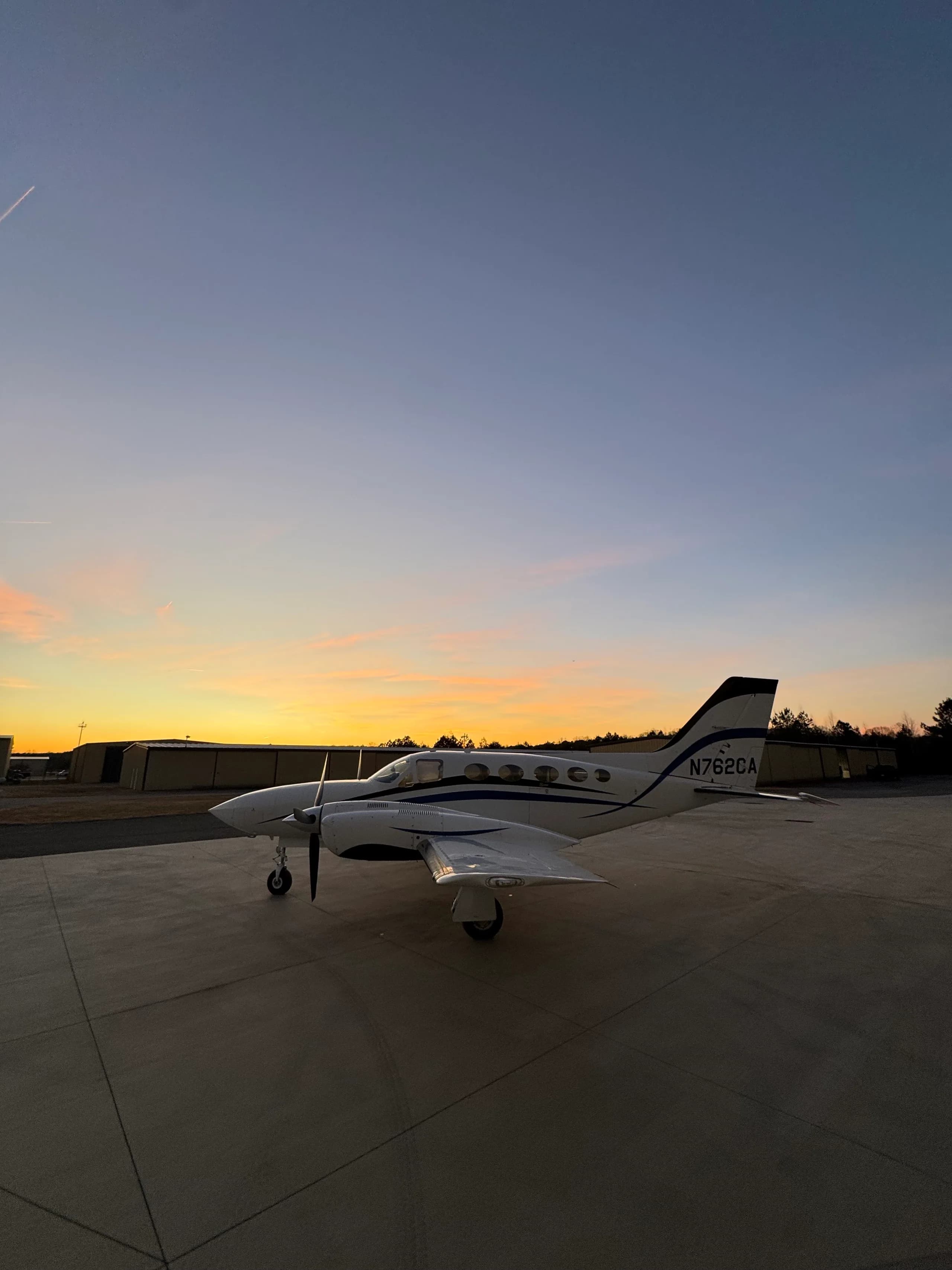 A white twin-engine aircraft with blue stripes, N762CA, parked on an airfield under a colorful sunset sky.