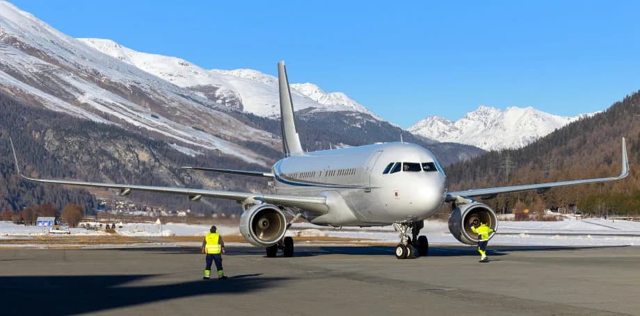 A silver Airbus A319 is parked on a snowy airport tarmac with ground crew, set against a backdrop of snow-capped mountains.