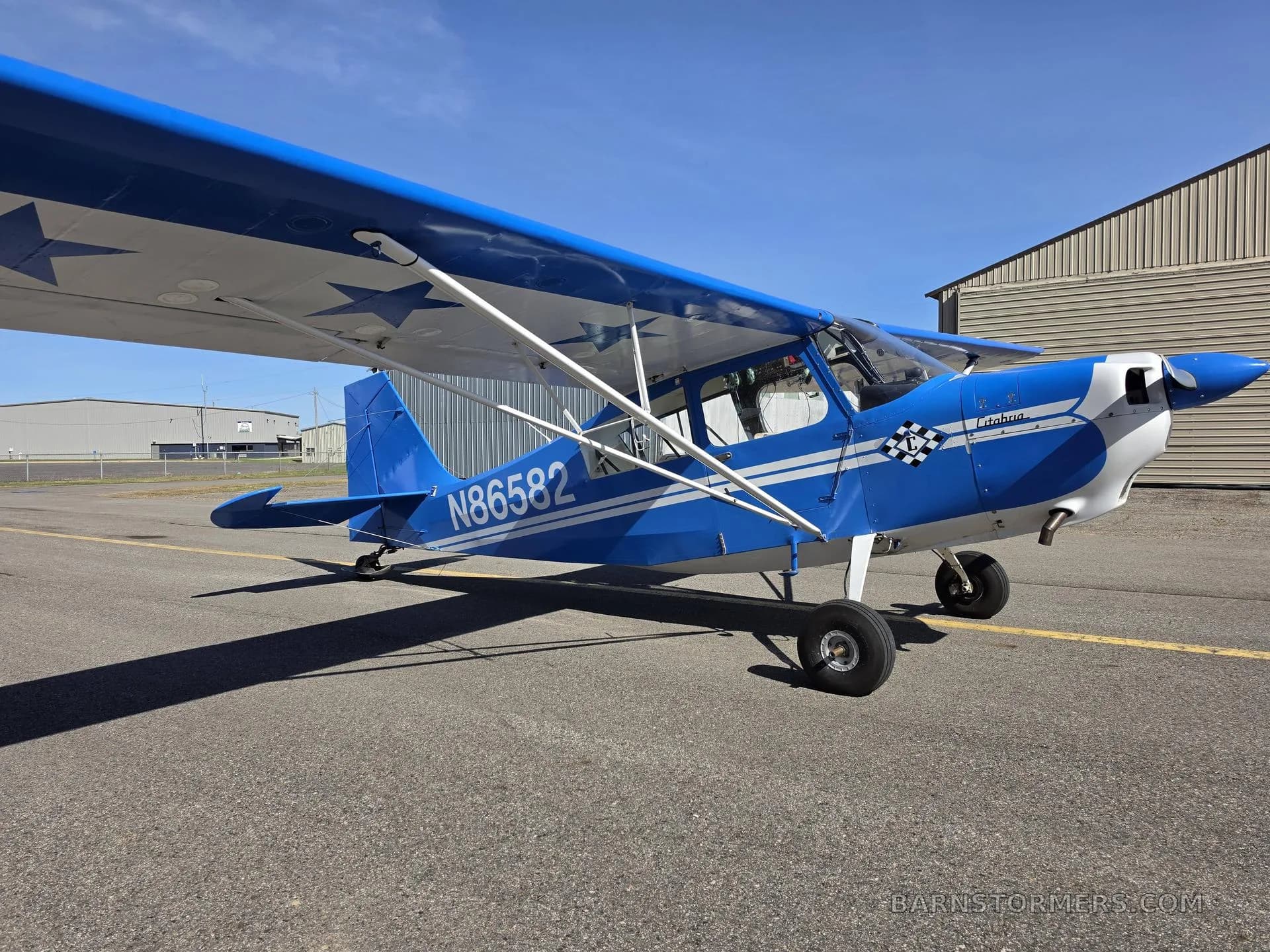 A blue and white single-engine propeller aircraft, tail number N86582, parked on an asphalt ramp with hangars under a clear sky.