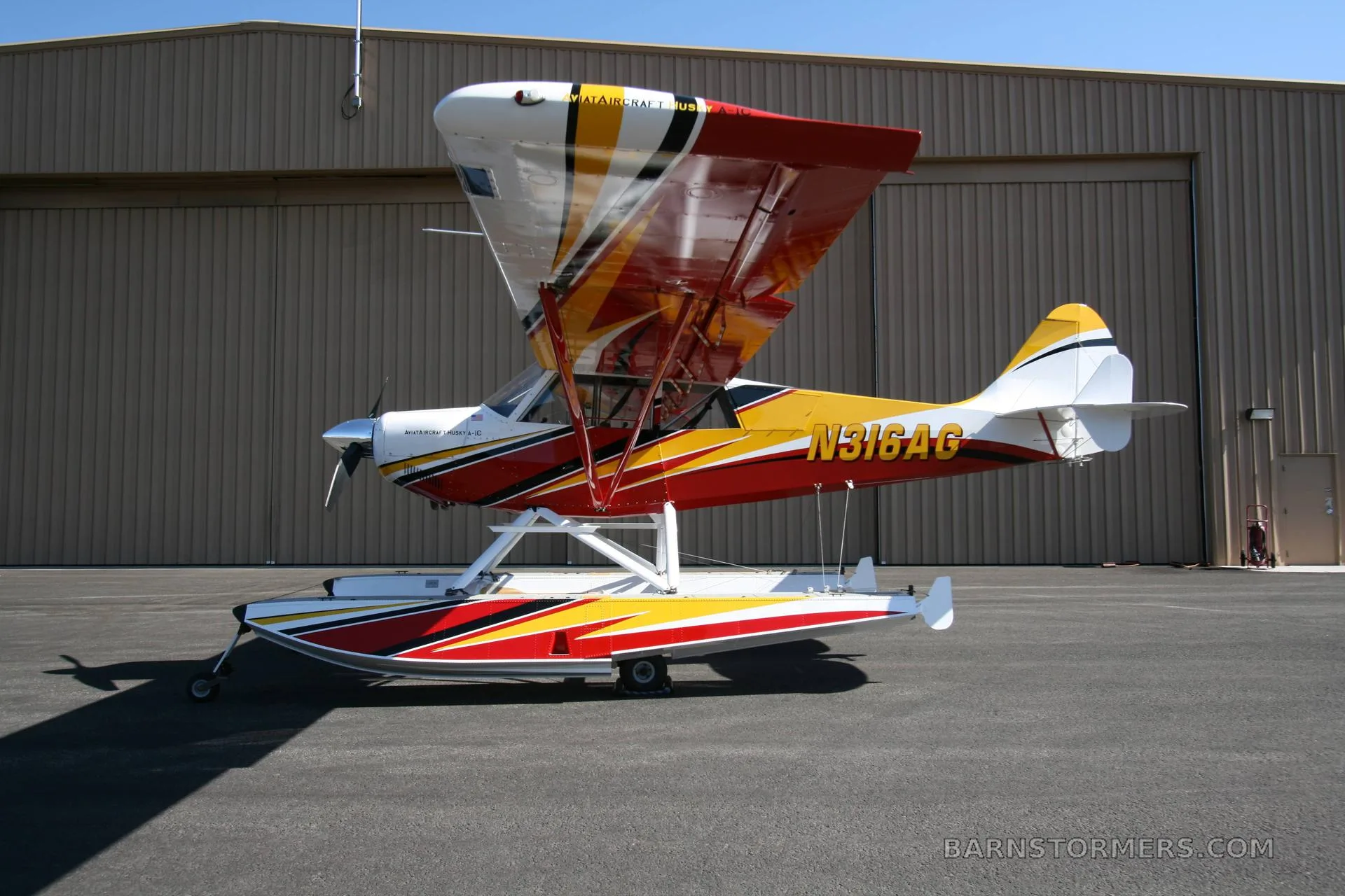 A red, yellow, and white floatplane with registration N316AG is parked on an asphalt tarmac in front of a hangar.