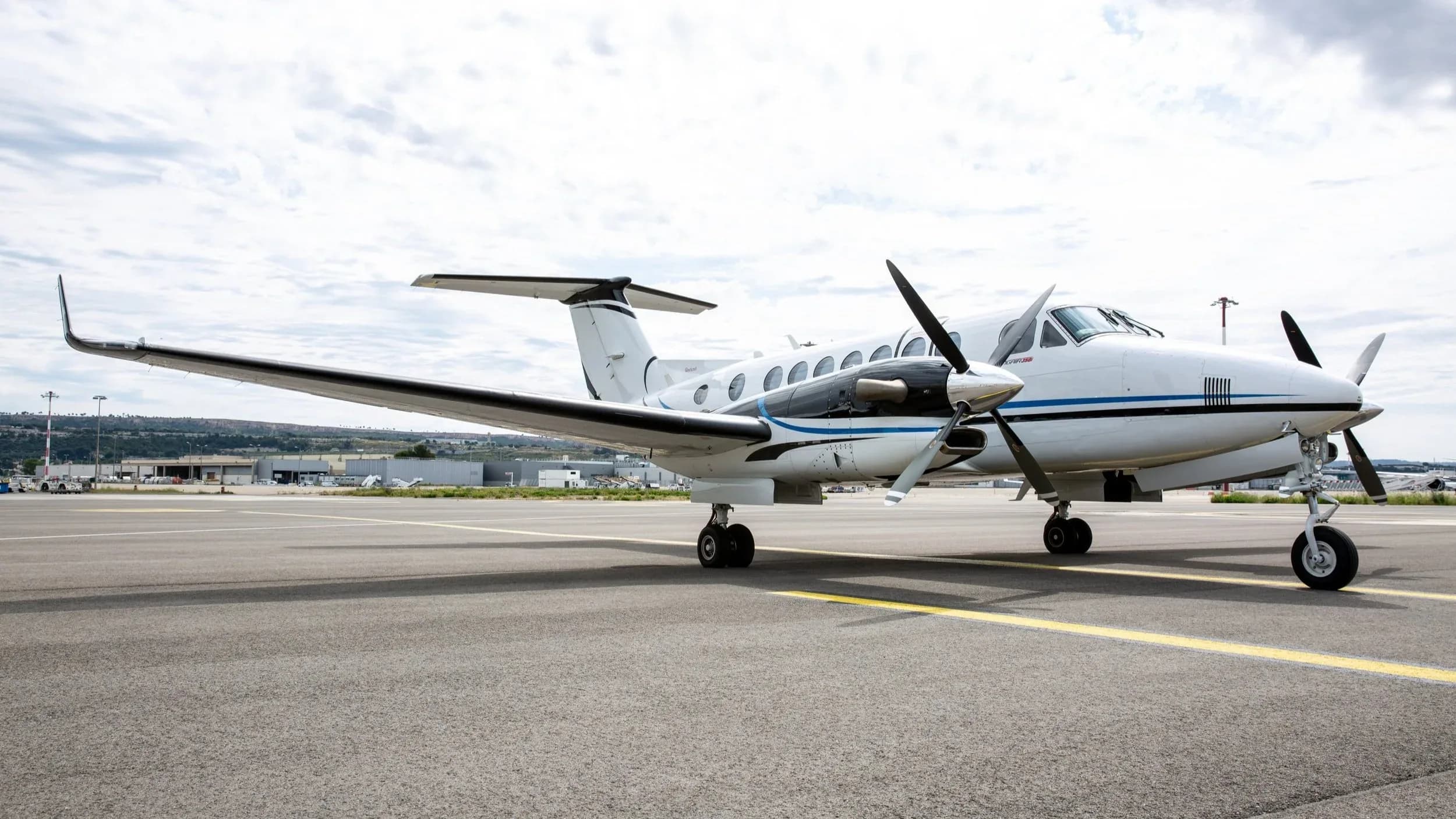 A white Beechcraft King Air 350i with black and blue accents is parked on an airport tarmac under a cloudy sky.
