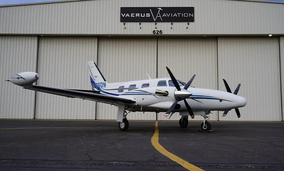 A Beechcraft Baron aircraft with a white and blue paint scheme is parked in front of a hangar.