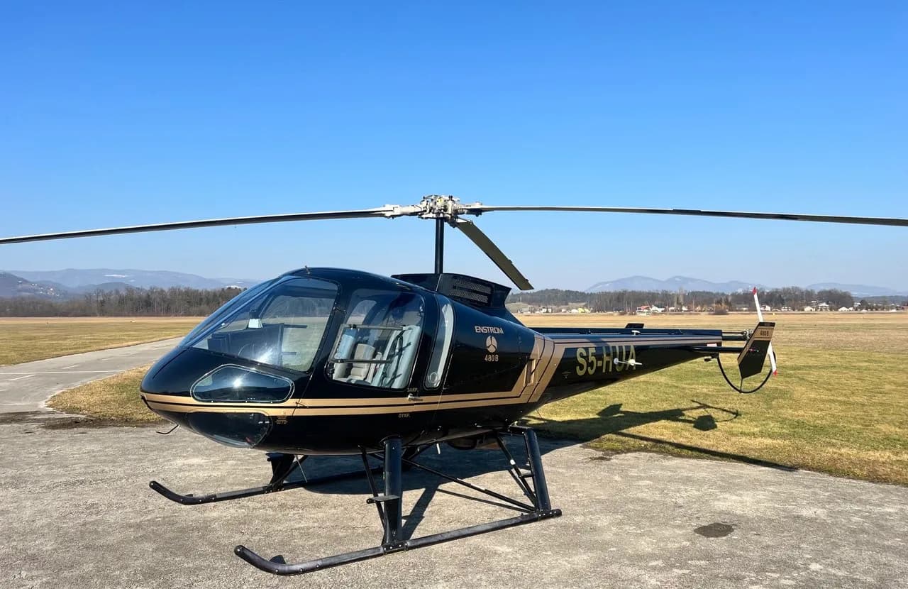 A black and gold Enstrom 480B helicopter (S5-HUA) parked on an airfield with mountains in the background.