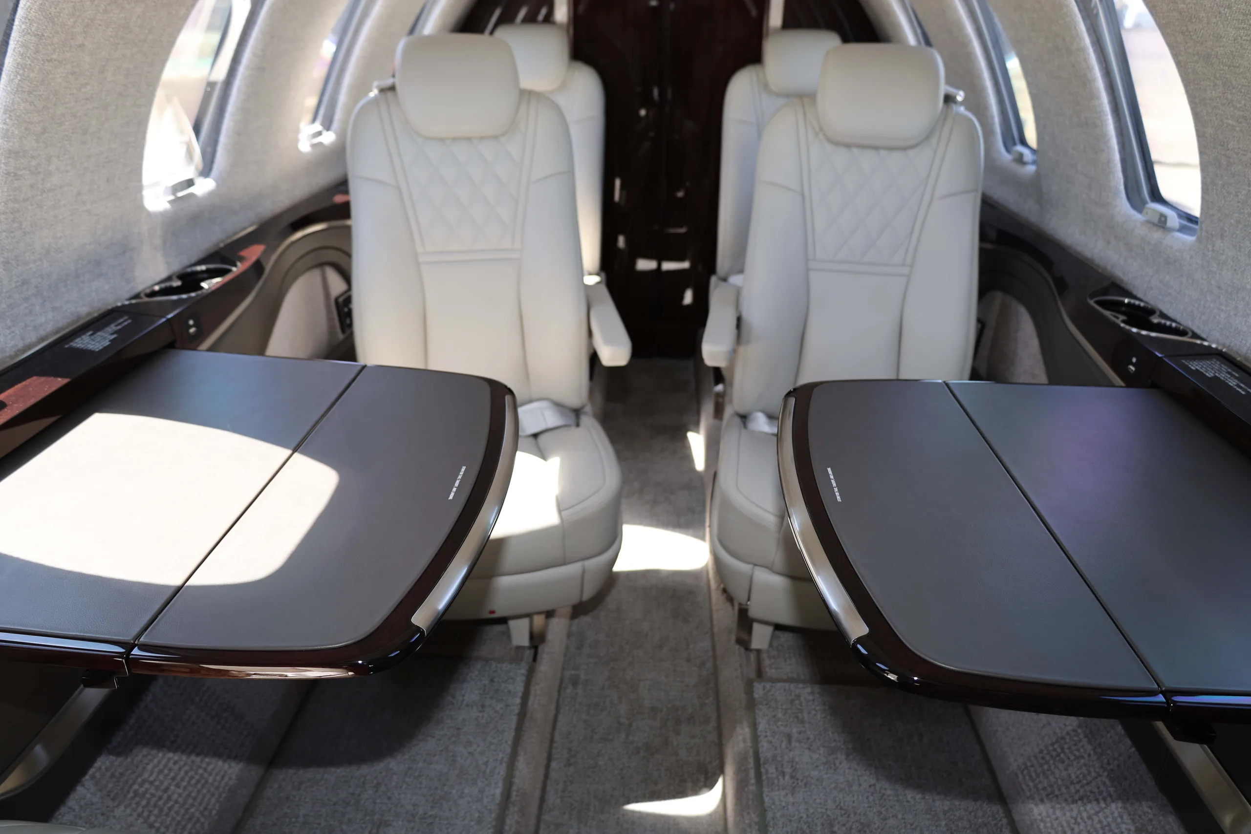 The interior cabin of a private jet, featuring two light-colored leather seats with their dark, fold-out tables extended.