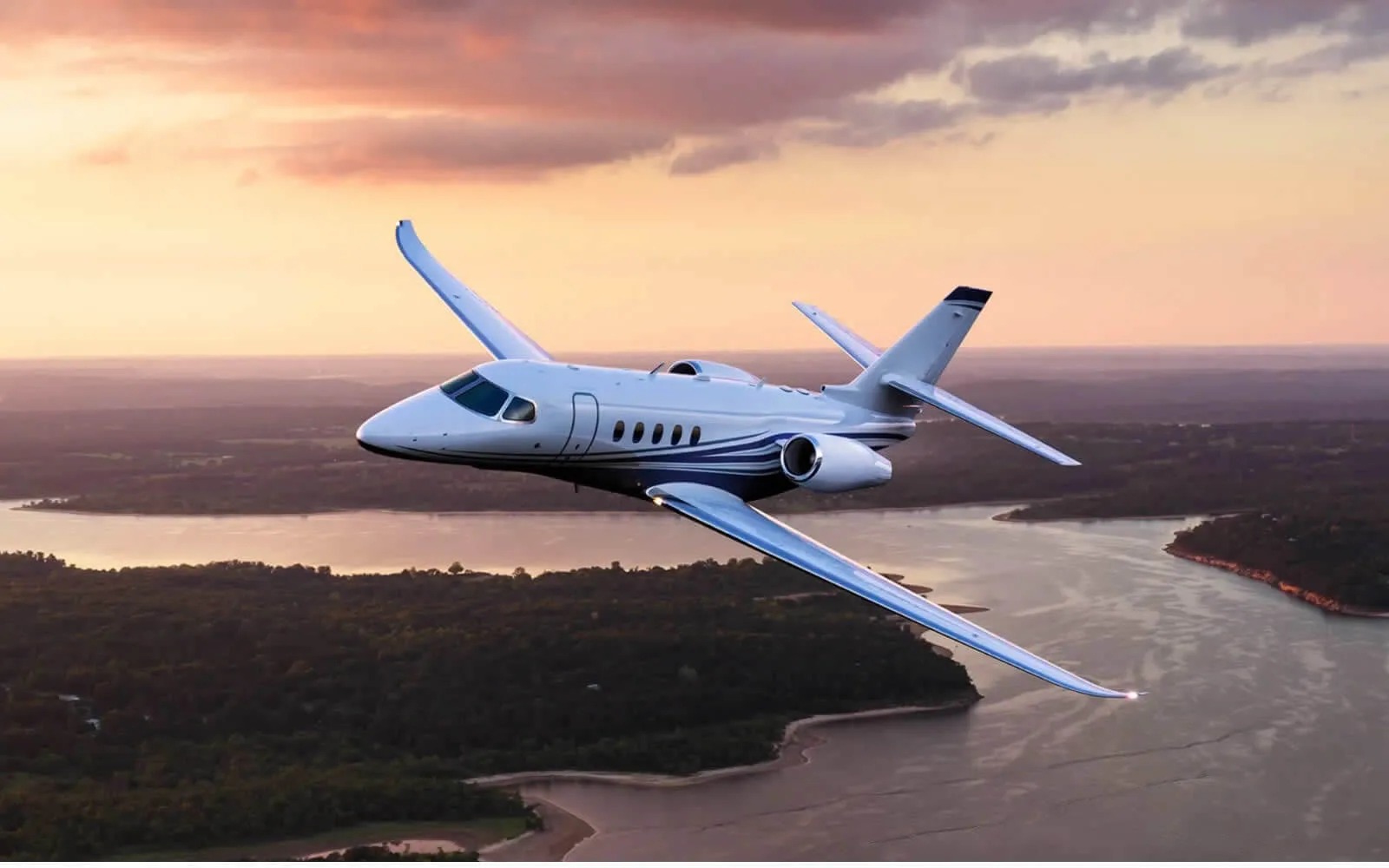 A white jet aircraft flies over a landscape at sunset.