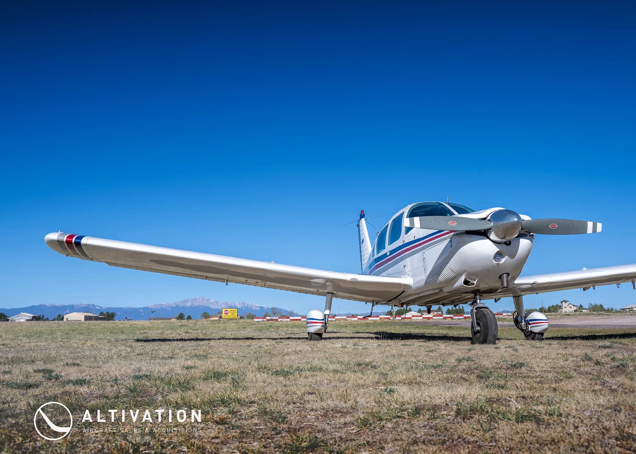 A low-angle view of a white Piper Cherokee aircraft with red and blue stripes on a grassy airfield.