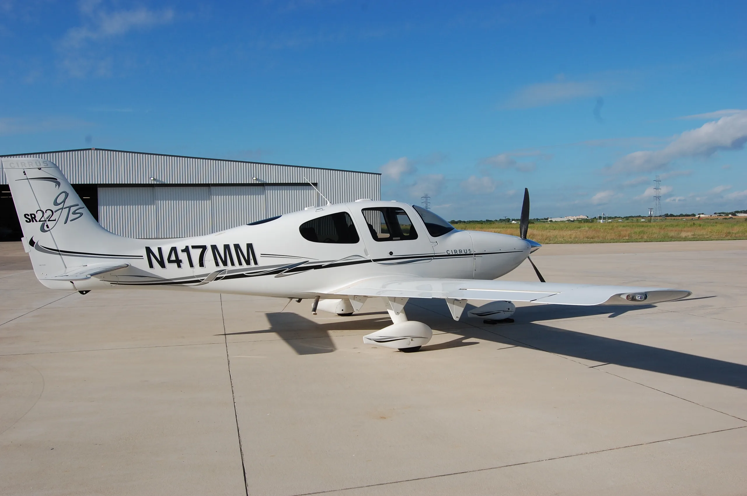 A three-quarter rear view of the white Cirrus SR22 aircraft, N417MM, parked on an airfield with hangars in the background.