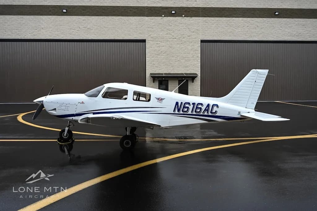 A white single-engine aircraft, N616AC, with blue stripes is seen from the side on a wet tarmac in front of hangars.