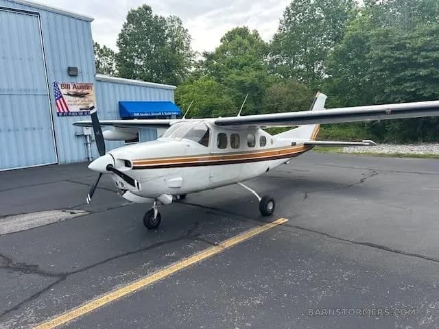 A white Cessna 337 Skymaster with brown and orange stripes is parked on an asphalt ramp next to a blue hangar.