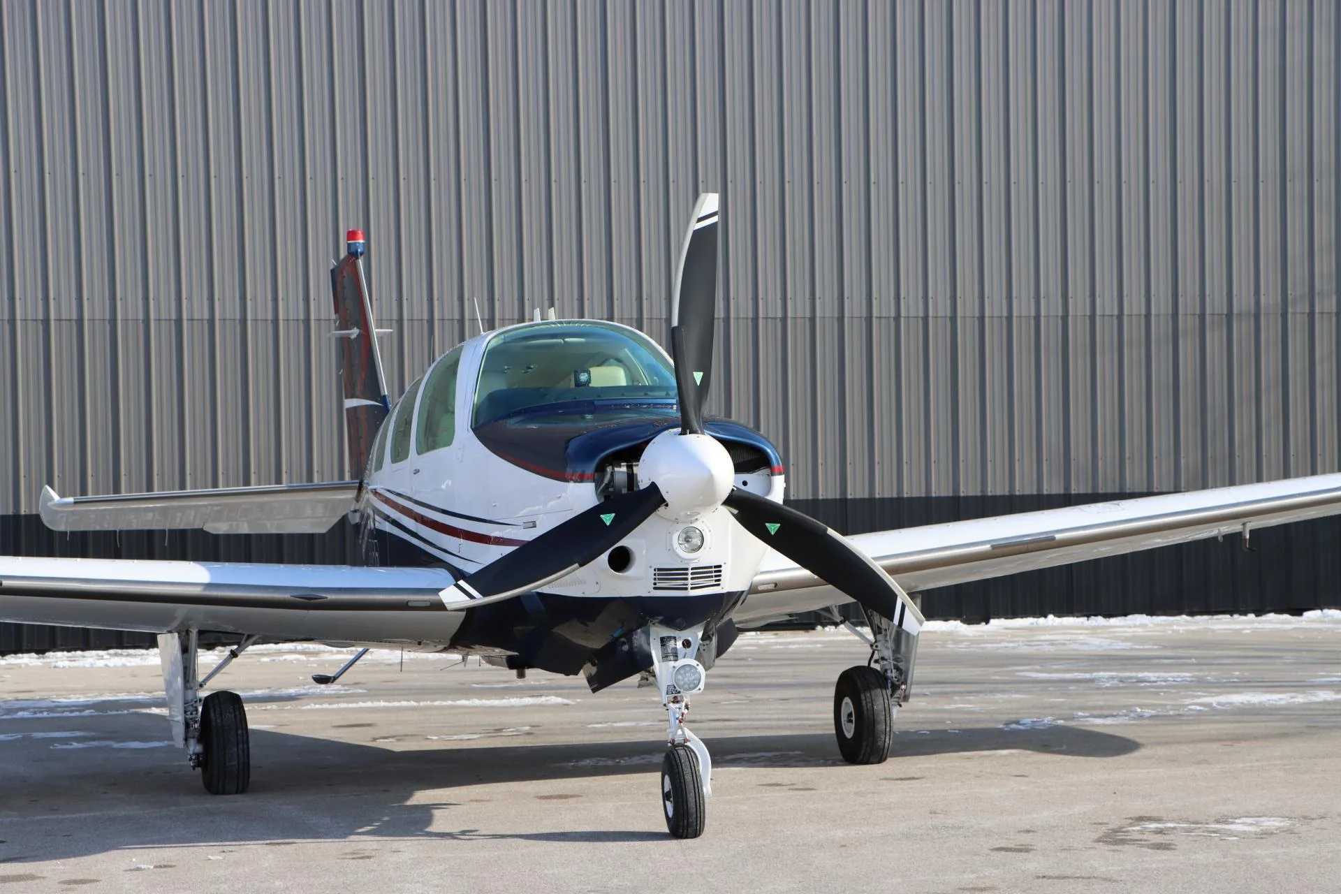 Close-up front view of a white, blue, and red single-engine aircraft with a three-blade propeller, parked on a partially snowy tarmac.