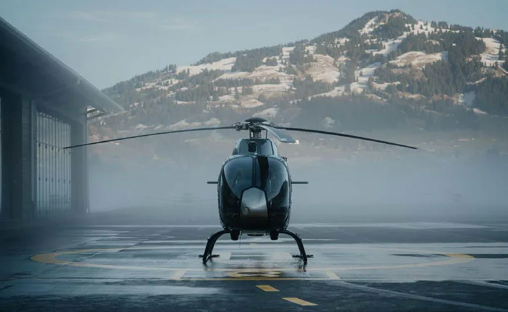 A dark grey helicopter is viewed from the front, sitting on a wet helipad with a snowy mountain in the background.