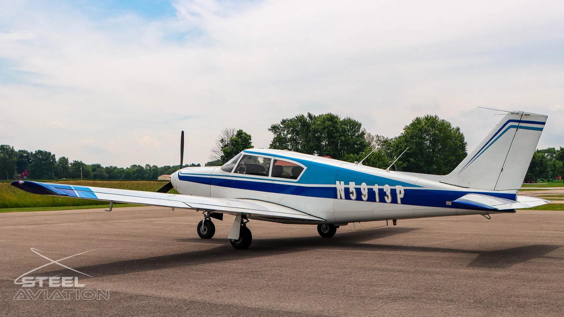 A side-rear view of the white and blue single-engine aircraft N5913P on a tarmac with trees in the background.