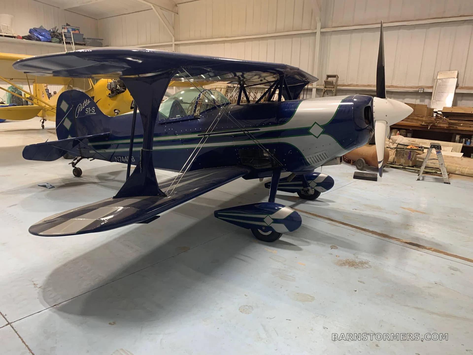 A blue and silver Pitts S1-S biplane with green stripes is parked inside a hangar.