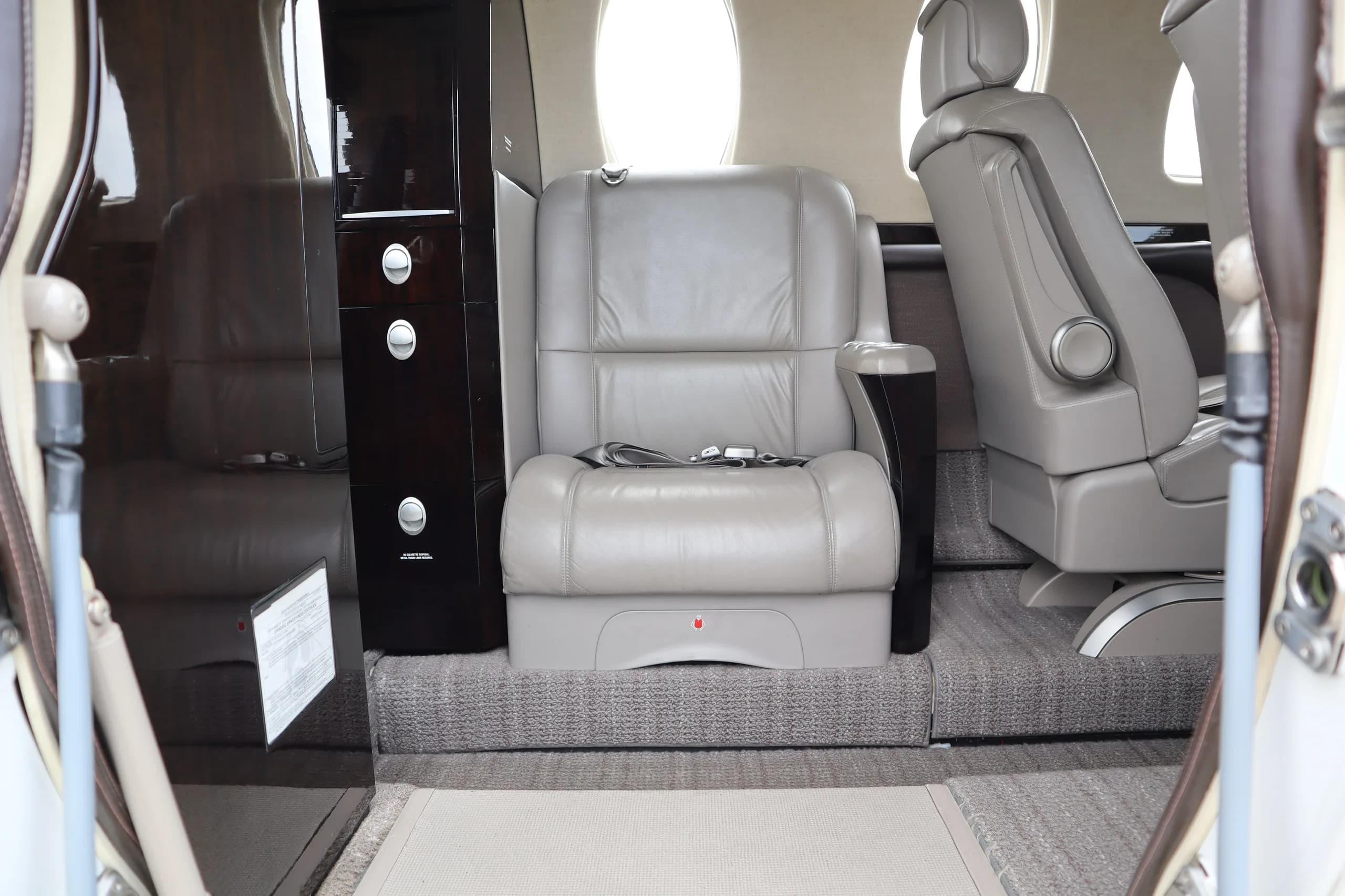 A view inside the business jet cabin showing a single grey leather seat next to a dark wood cabinet.