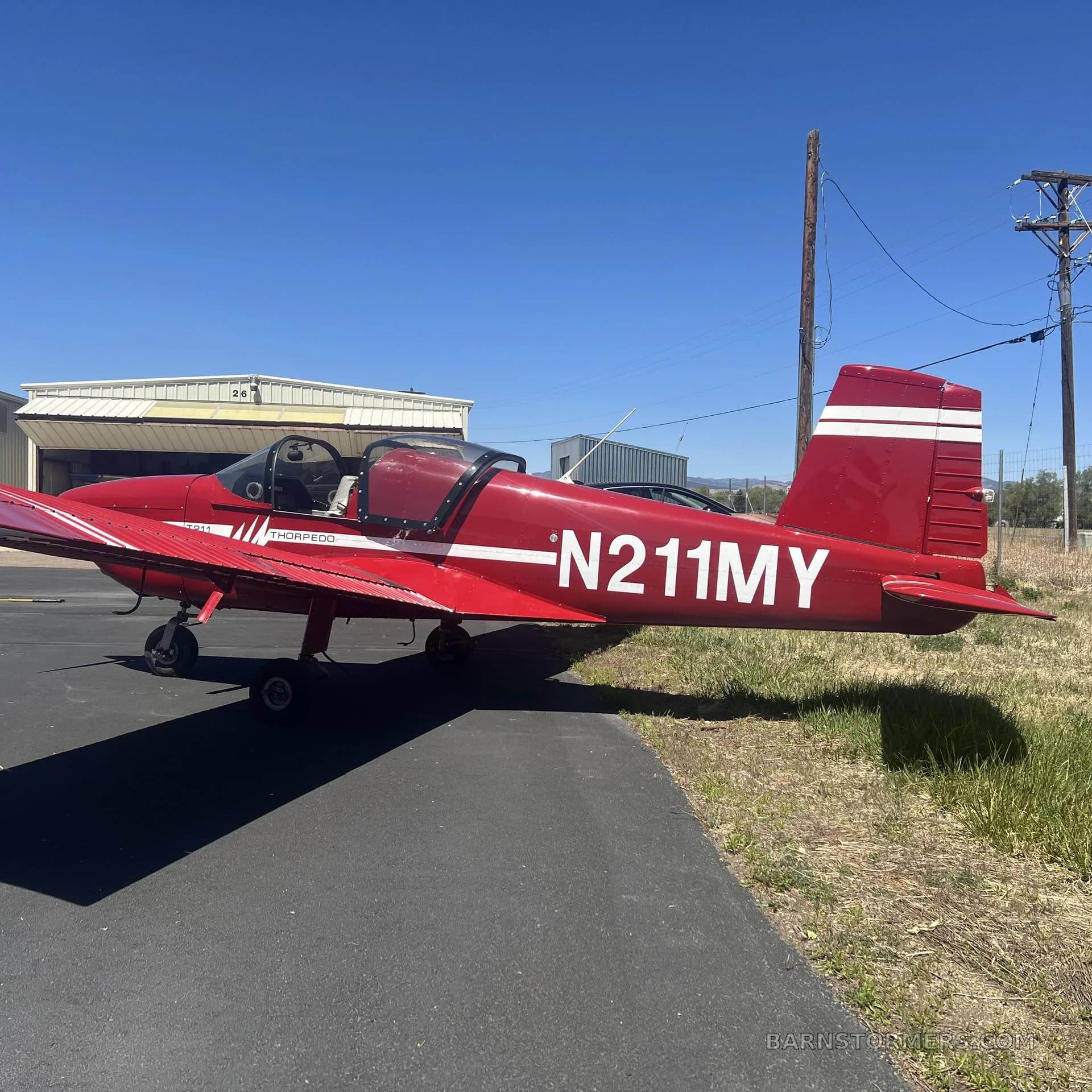 A red Thorp T211 Torpedo aircraft with white stripes and registration N211MY, parked on an asphalt taxiway next to a hangar.