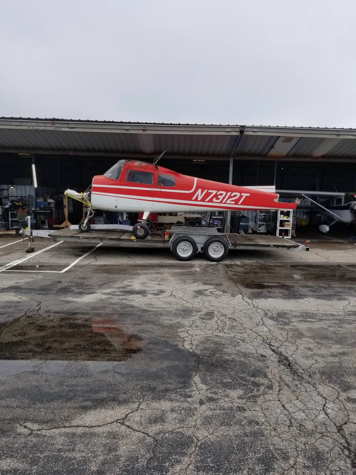 A red and white single-engine aircraft, N7312T, with its engine cowling removed, sits on a trailer outside a hangar on a wet tarmac.