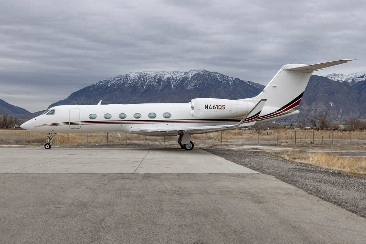 A white Gulfstream business jet with red and black stripes (N461QS) parked on a concrete pad with snowy mountains in the background.
