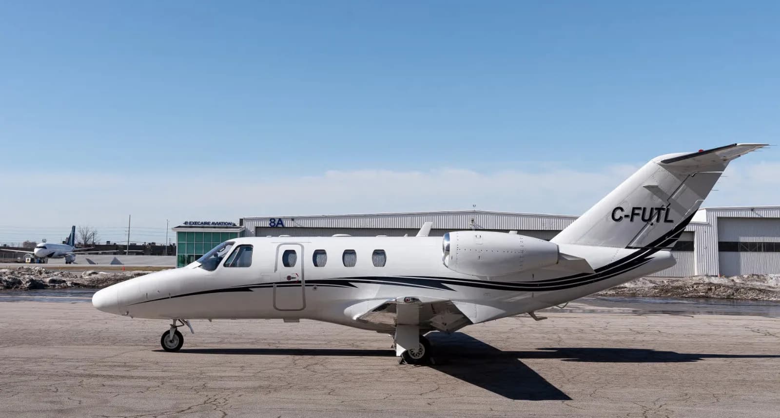 A white Cessna Citation CJ3 is parked on the tarmac with a hangar in the background.