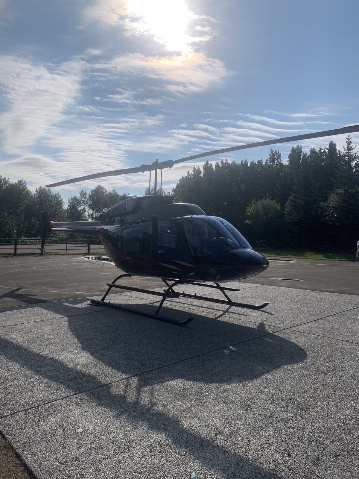 A black and red Bell 407 helicopter is parked on a concrete pad on a sunny day.