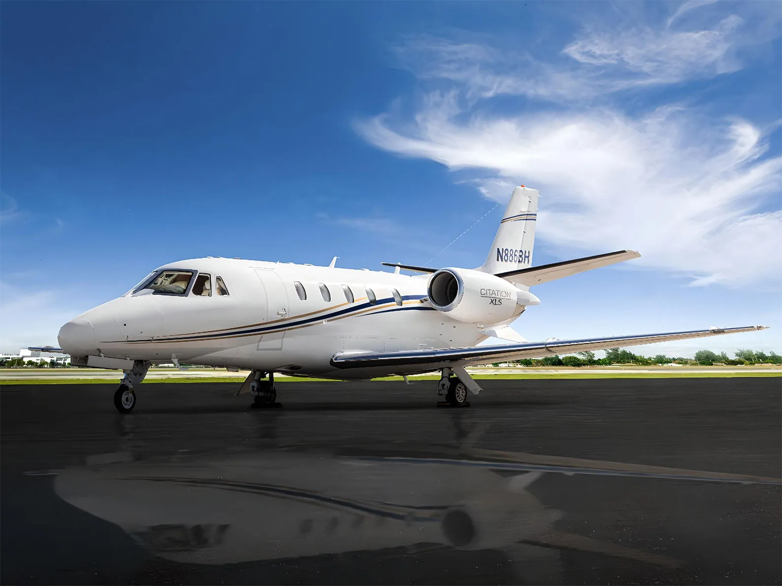 A white Cessna Citation XLS private jet with blue and gold stripes parked on a wet tarmac under a dramatic sky.