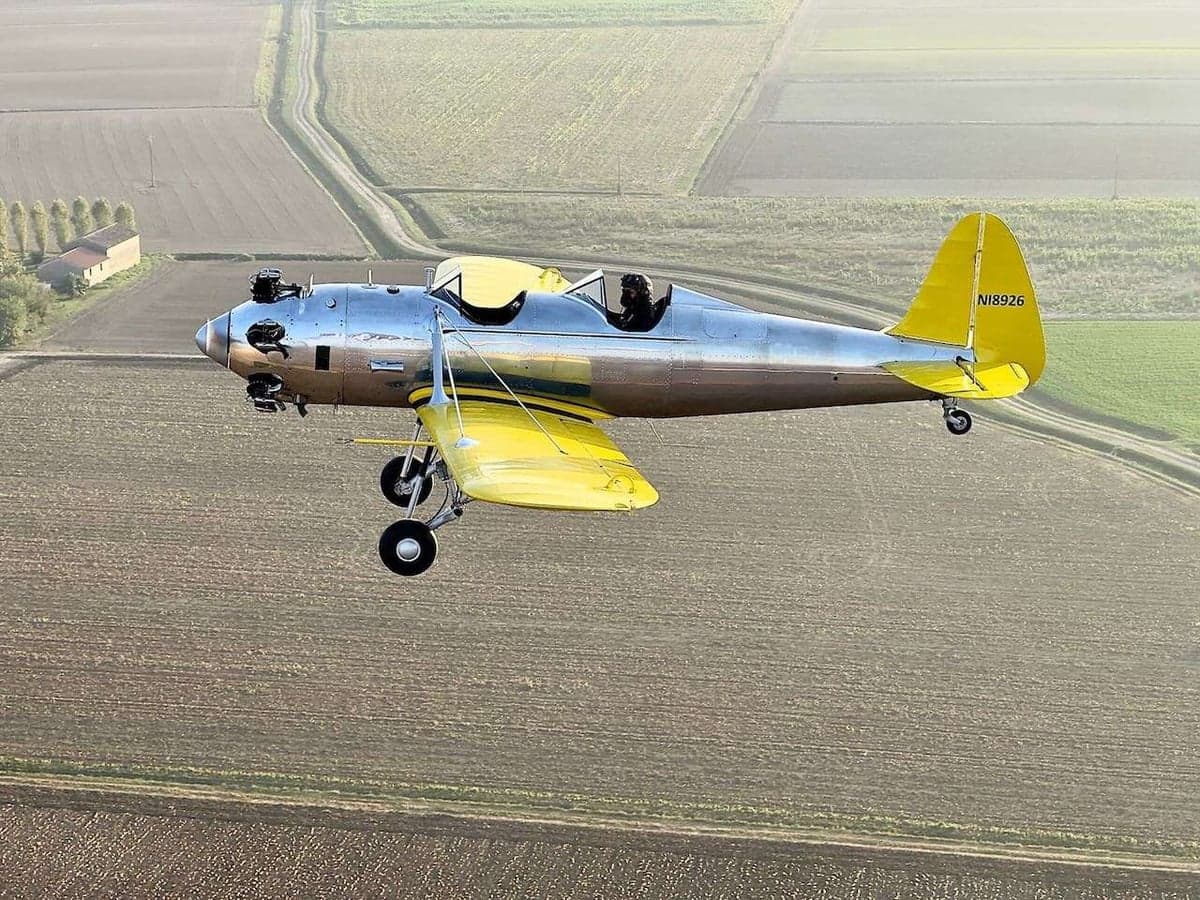 A silver and yellow aircraft, registration N18926, in flight over a rural landscape.