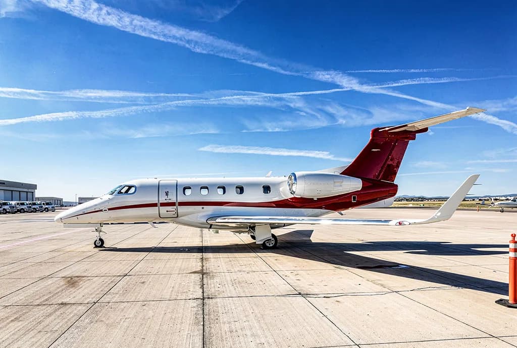 A white and red Embraer Phenom 300 private jet parked on an airport tarmac with contrails in the sky.