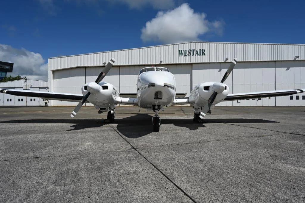 A white twin-engine propeller aircraft with black propellers, viewed from the front, parked on a concrete apron in front of a white "WESTAIR" hangar under a blue sky.