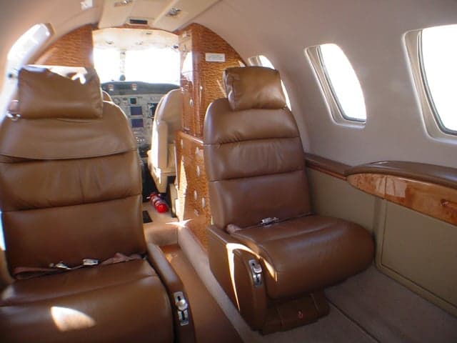 The interior cabin of a private jet with brown leather seats and wood paneling, looking towards the cockpit.