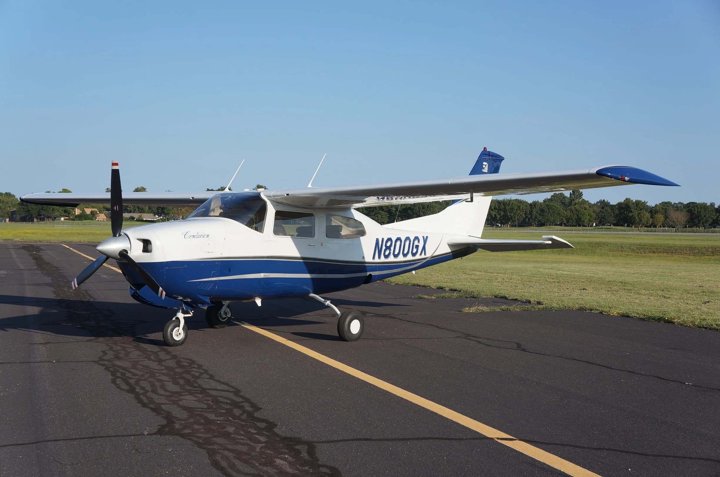 A white and blue Cessna 206 Stationair aircraft, N800GX, is parked on an airport tarmac with grass and trees in the background.