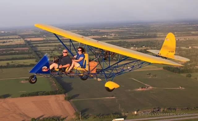 A yellow and blue two-seat ultralight aircraft with two occupants flying over green and brown fields.