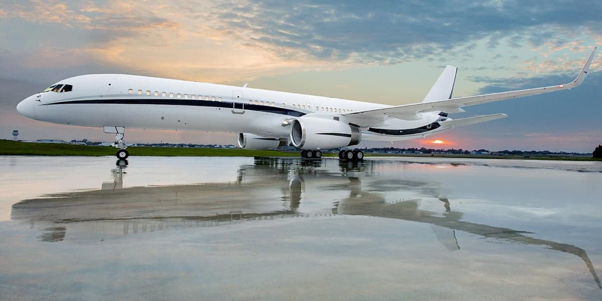 A white and navy Boeing 757 business jet is parked on the tarmac at sunset, reflected in the wet surface.