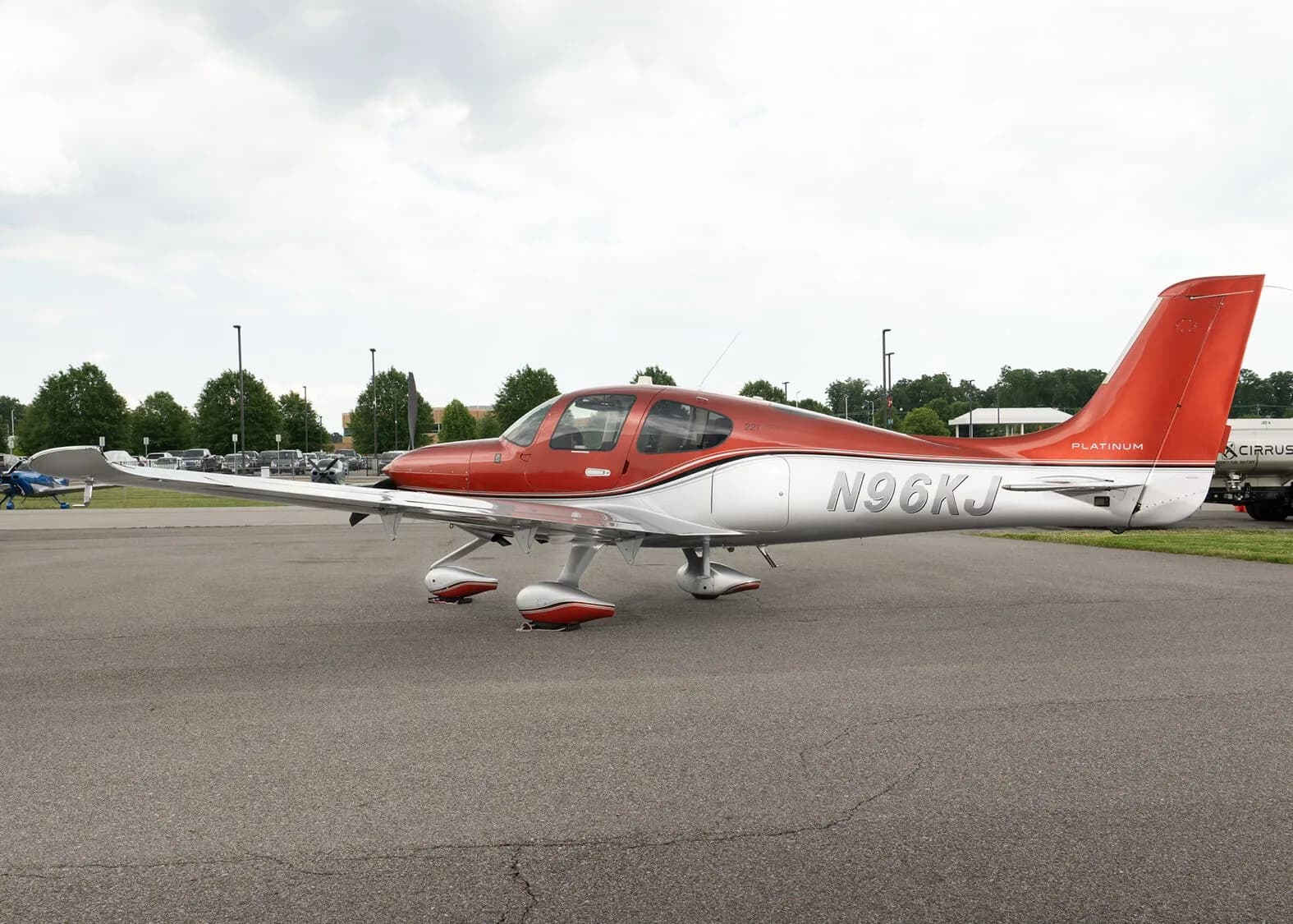 A red and silver Cirrus SR22T aircraft, registration N96KJ, parked on an asphalt apron with other aircraft and trees in the background.
