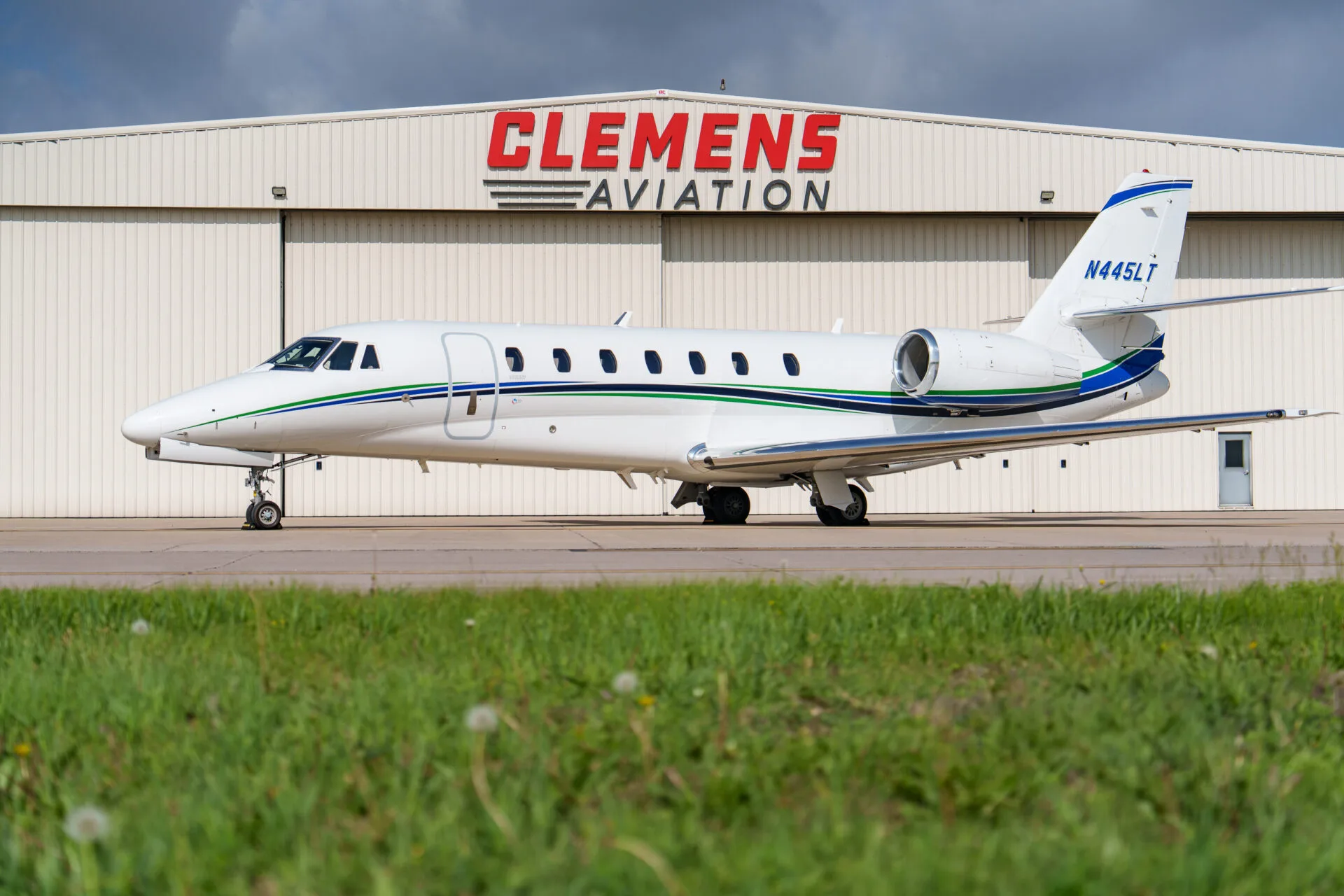 A white Cessna Citation business jet with green and blue stripes parked on an airfield with grass in the foreground.