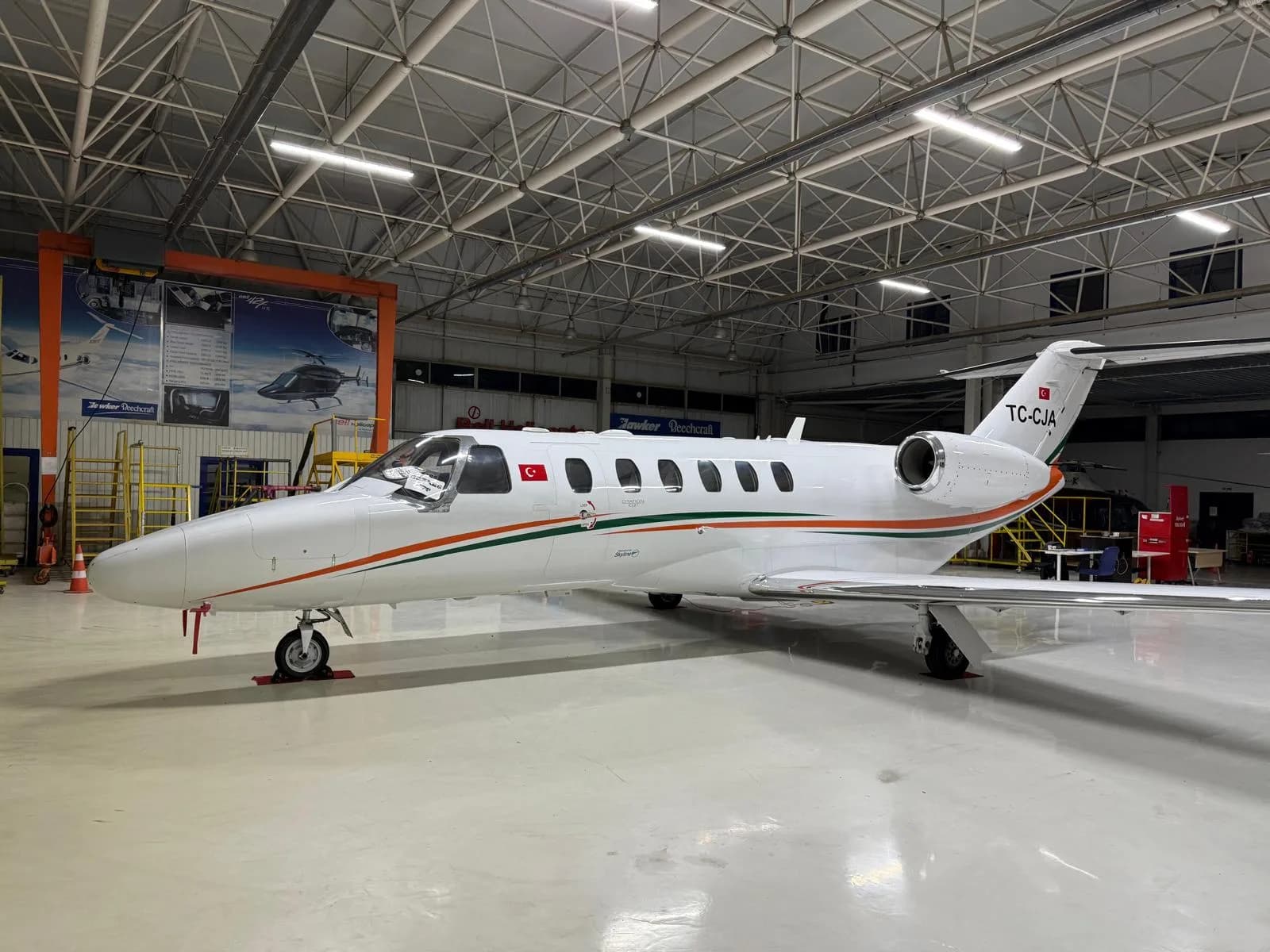 A white business jet with orange and green stripes and Turkish flag tail number TC-CJA, parked in a hangar.
