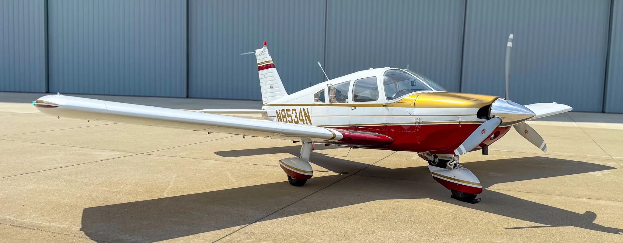 A white, red, and gold Piper Cherokee aircraft (N8534N) parked on an airfield in front of a hangar.