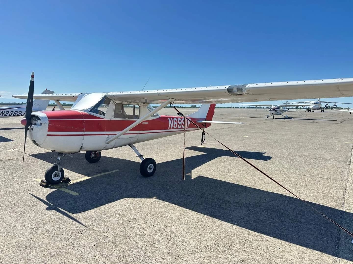 A red and white Cessna 150, tail number N6991F, parked on an asphalt apron with other aircraft in the background.