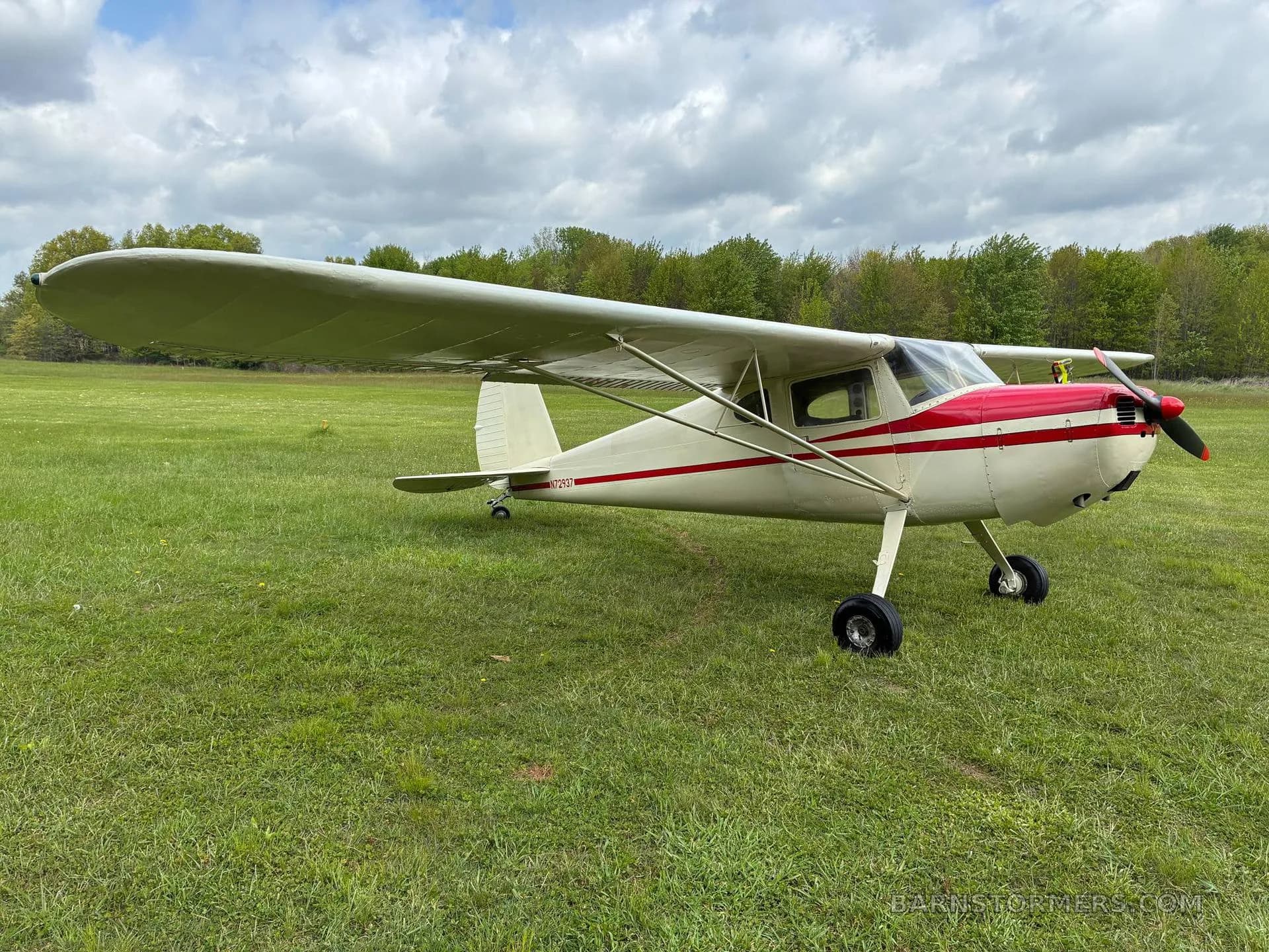A white and red single-engine high-wing aircraft with taildragger landing gear is parked on a grassy field under a cloudy sky.