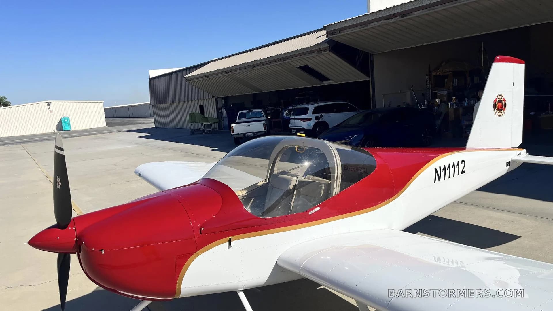 A red and white single-engine aircraft with registration N11112 is parked on an airfield apron in front of an open hangar.
