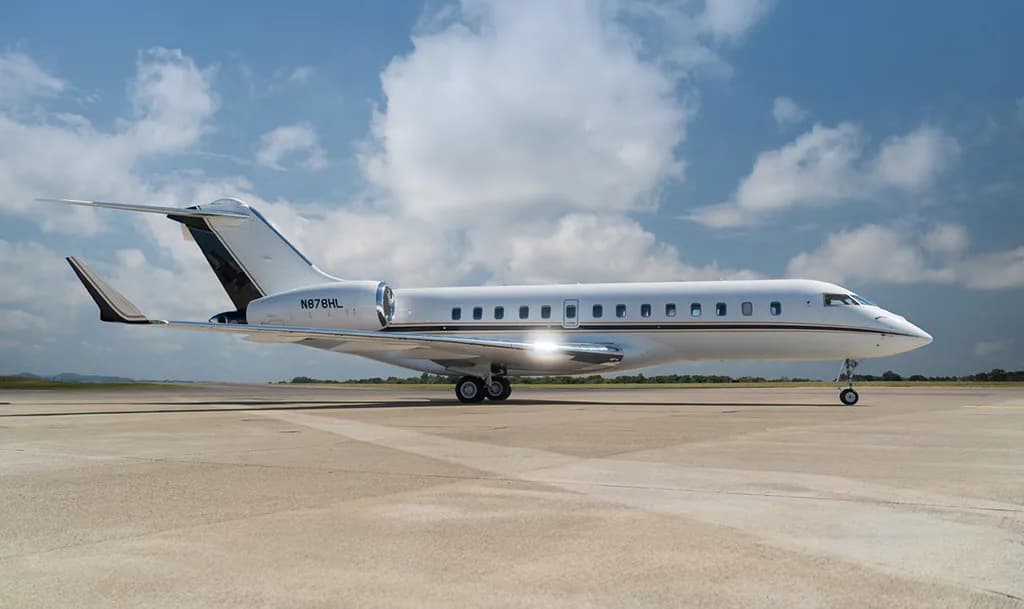 A white Bombardier Global 6000 private jet with black and gold accents on the tail, registration N878HL, parked on an tarmac under a partly cloudy sky.