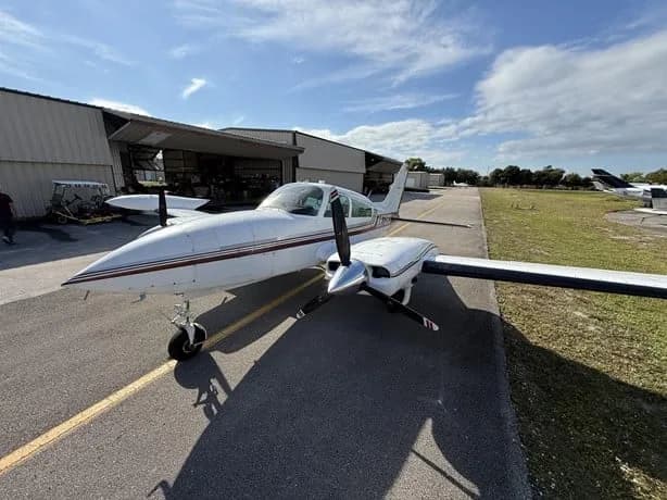 A white twin-engine aircraft with red and black stripes, viewed from the front-side on an airfield.